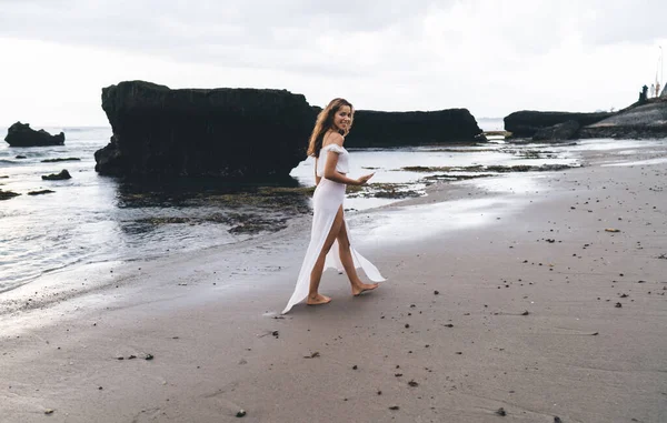 Deniz kıyısında akıllı telefondan mesajlaşırken gülümseyen ve kameraya bakan beyaz elbiseli, çıplak ayaklı, iyimser bir bayan gezgin.