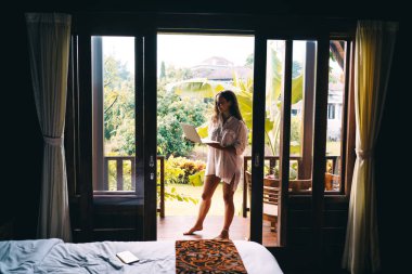 Modern yatak odasının balkonunda güneşli bir yaz gününde çıplak ayakla dizüstü bilgisayar kullanan beyaz tişörtlü genç bir kadın.