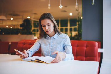 Modern kafeteryadaki açık günlüklü ve cep telefonlu, uzun saçlı, rahat gömlekli bir kadın.