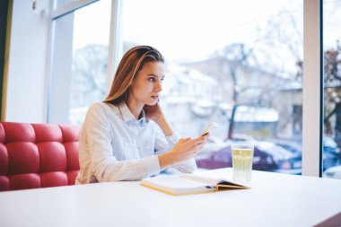 Açık not defteri ve bir bardak su sörfü akıllı telefonuyla masada otururken gözünü kaçıran odaklanmış kız öğrenci.