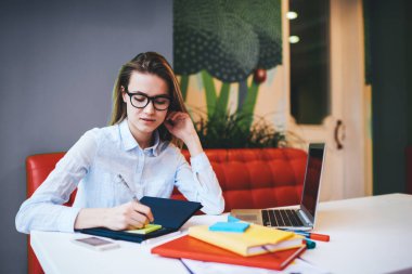 Genç, konsantre olmuş, günlük giysiler içinde dizüstü bilgisayar ve not defterleriyle masada oturmuş proje üzerinde çalışırken notlar alan kadın bir serbest yazar.