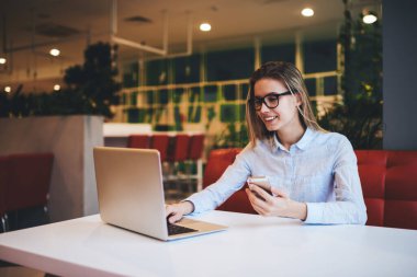 Gözlüklü ve açık renk tişörtlü neşeli kadın netbook ile masada oturuyor ve gündüz kafede çalışırken akıllı telefon kullanıyor.