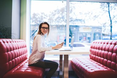 Günlük kıyafetlerle gülümseyen serbest çalışan, netbook ile masaya oturan ve gündüz kafede proje üzerinde çalışırken akıllı telefon kullanan kadın.