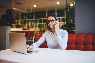 Gözlüklü ve beyaz tişörtlü gülümseyen kadın girişimci akıllı telefonla konuşurken dizüstü bilgisayarla proje üzerinde çalışıyor ve gündüzleri kafede başka yere bakıyor.