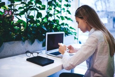 Gündelik kıyafetli genç bir kadın, açık modern bir kafede çalışırken dizüstü bilgisayar ve defterle masada oturup akıllı telefonları karıştırıyor.