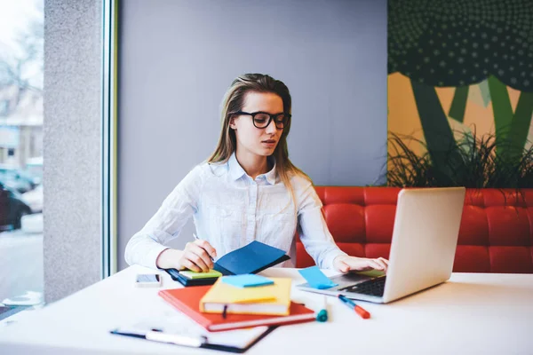 Gündelik kıyafetli, elinde defterlerle masada oturan ve modern iş yerinde uzaktan çalışırken dizüstü bilgisayar kullanan ciddi bir kadın serbest yazar.