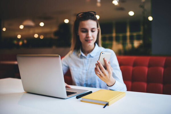 Young glad female remote worker in casual clothes with eyewear sitting at table with laptop and reading messages on smartphone