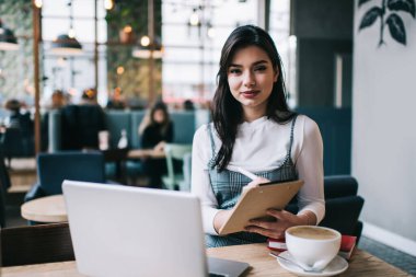 Bir fincan kahveyle masaya oturup not alırken kameraya bakan mutlu kadın uzaktan çalışan.