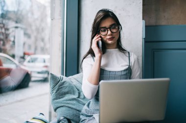 Ciddi kadın uzaktan çalışan, pencerenin yanında dizüstü bilgisayarla oturuyor ve hafif çalışma alanında çalışırken cep telefonuyla sohbet ediyor.
