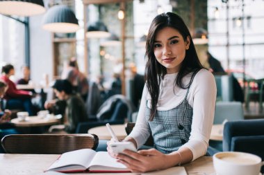 Uzun siyah saçlı, sırıtan bir kız öğrenci kameraya bakarken masada akıllı telefonuyla oturmuş, defterine notlar yazıyormuş.