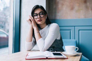 Kafede, kafeteryada, elinde kahve ve defterle oturup akıllı telefondan müzik dinleyen ve gözlerini kaçıran düşünceli bir kadın.