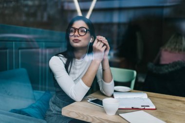 Kendine güvenen koyu renk saçlı kadın penceresinden. Günlük kıyafetler ve gözlüklü. Elinde kahve fincanı ve not defteriyle oturmuş akıllı telefondan müzik dinliyor.