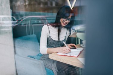 Uzun saçlı, zarif giyinmiş, gözlüklü bir kadının penceresinden not defterine yazarken, kafeden uzak bir iş için akıllı telefon kullanıyor.