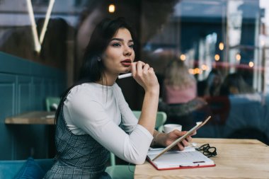 Kafede notlarla çalışırken şık elbiseli, odaklanmış koyu renk saçlı bir kadının camdan bakışları.