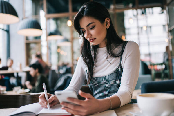 Pensive female student using mobile phone and writing notes in notepad while sitting at table in light cafe and doing task