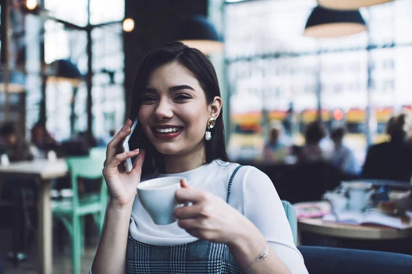 コーヒーのマグカップを持つ陽気な女性離れて見ている間電話会話で現代のカフェテリアでぼやけた背景に休憩中に — ストック写真