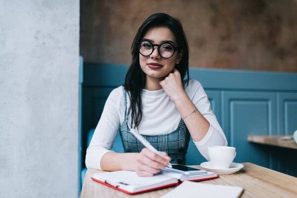 Content woman in casual wear and eyeglasses looking at camera and writing in notebook while working remotely with smartphone in cafe