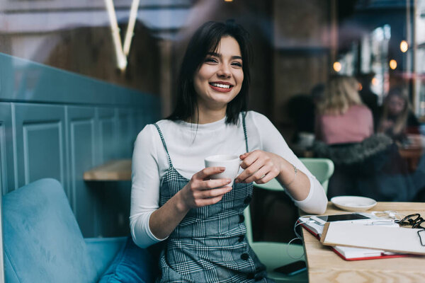 Happy young female student wearing trendy clothes sitting at table with notebook and papers and drinking hot beverage while preparing for exams in cafe