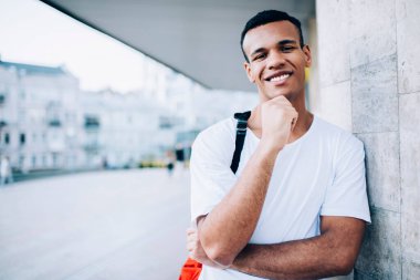 Tişörtünde dişlek bir gülümseme ve çenesine bakan genç, pozitif siyah bir erkek sokakta dururken kameraya bakıyor.