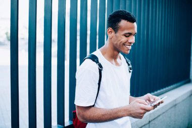 Sırt çantasıyla sokakta metal çitlerin yanında durup cep telefonuyla oyun oynayan neşeli Afro-Amerikan erkek.