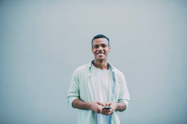 Beyaz tişörtlü, beyaz tişörtlü, beyaz arka planda tek başına kameraya bakarken akıllı telefon kullanan gülümseyen genç bir zenci.