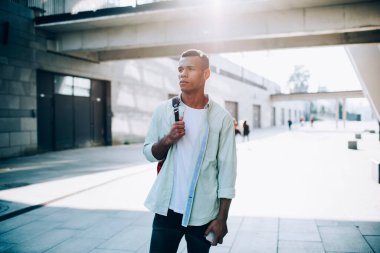 Düşünceli genç siyah bir adam. Sırt çantalı, binaların yakınındaki caddede geziniyor ve güneşli yaz günlerinde cep telefonuyla köprüye bakıyor.