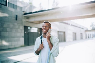Gündelik giysiler içinde mutlu genç bir adam inşaat ve köprü yakınında geziniyor. Akıllı telefonla konuşuyor ve güneşli yaz günlerinde kameraya bakıyor.