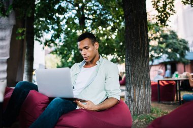 Ciddi genç siyahi bir adam günlük kıyafetleriyle şezlongda oturuyor ve yazın ağaçların yanında netbook 'ta çalışıyor.