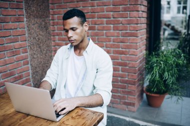 Ciddi, genç, siyah, uzaktan kumandalı bir işçi. Gündüz vakti masa başında oturmuş çalışma alanındaki tuğla duvarın yanındaki yeşil fabrikanın yanında Netbook kullanıyor.