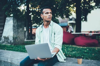 Konsantre olmuş genç Afro-Amerikan bir adam. Gündelik giysiler içinde. Laptop üzerinde çalışıyor. Yaz akşamları şehir parkında oturuyor ve gözlerini kaçırıyor.