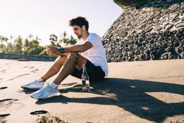 Spor giyim ve kol saati giymiş sakallı bir erkeğin kumsalda yazın elinde bir şişe suyla oturup kulaklıkla müzik dinlemesinin yan görüntüsü.