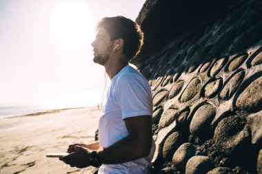 Elinde cep telefonu olan, kulaklıkla müzik dinleyen ve sahile yakın kumsalda duran düşünceli sakallı bir erkeğin yan görüntüsü.