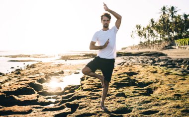 Sırtında uzun sakallı bir erkek, günlük yaz kıyafetleri giyiyor. Tropikal bir tatil köyünde güneşin doğuşu sırasında kumlu sahil kenarında kollarıyla dengede duruyor.