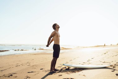 Sörf tahtasının yanında kumlu sahilde duran ve deniz dalgalarında sörf yapmadan önce kollarını esneten kaslı erkek sörfçü.