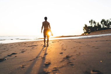 Deniz kenarlarında sörf tahtası ve renkli günbatımında ayak izleri olan çıplak gövdesiyle okyanusa hayran olan tanınmamış erkek sporcuların arka planı.