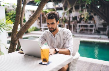Çizgili tişörtlü, otelin bölgesinde otururken ve dizüstü bilgisayar kullanırken internetten bilgi arayan konsantre olmuş, yakışıklı, sakallı adam.
