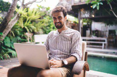 Çizgili gömlekli, sakallı, koltukta oturmuş, parlak dişli tebessümle kameraya bakarken interneti taramak için dizüstü bilgisayarı kullanan mutlu genç adam.