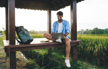 Neşeli, traşsız, uzaktan kumandalı bir çalışan netbook 'ta internette sörf yaparken güneşli bir günde kırsalda sırt çantasının yanında yerde oturuyor.