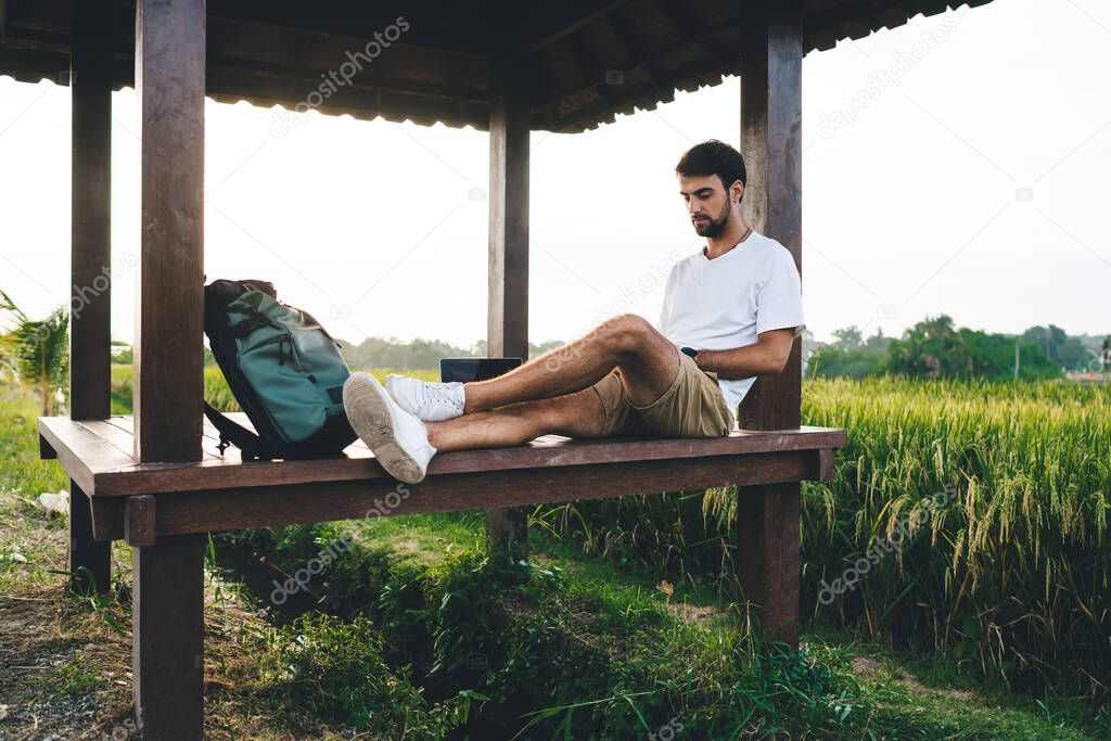 Young attentive male traveler sitting with crossed legs on floor of wooden alcove with rucksack while looking down against lush rice field in countryside