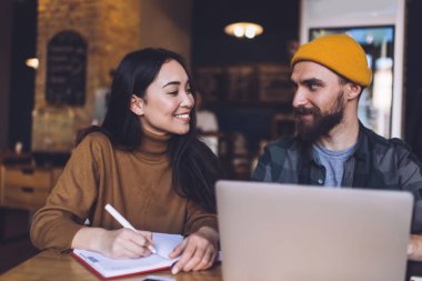 Şık şapkalı genç sakallı adam dizüstü bilgisayarda çalışıyor ve not defterine kalemle not alıp gülümseyen genç iş arkadaşına bakıyor.