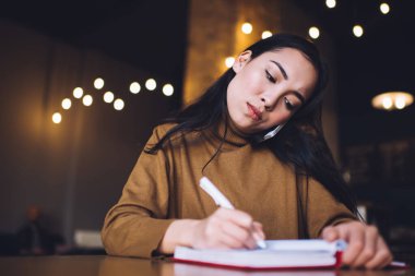 Düşük açılı, eğik kafalı, balıkçı yakalı, akıllı telefondan insanları dinleyen ve kafede otururken notlar alan esmer bir kız.