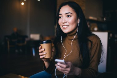 Bir fincan sıcak içecekle gündelik giysiler içinde gülümseyen genç bir kadın kafede otururken cep telefonundan müzik dinliyor.