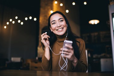 Gündelik giysiler içinde gülümseyen genç bir kadın karanlık kafede otururken cep telefonu kullanarak neşeli bir şekilde müzik dinliyor.