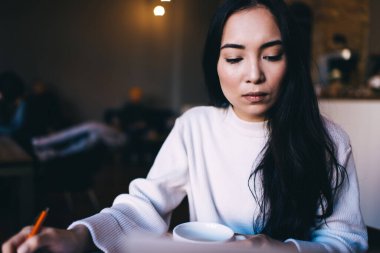 Odaklanmış beyaz kazaklı bir kadın, elinde bir fincan sıcak içecekle kafe 'nin karanlık köşesindeki masada otururken evrak işleriyle uğraşıyor.