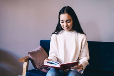 Gündelik kot pantolon ve süveter giyen düşünceli genç bayan öğrenci modern dairede rahat bir kanepede otururken ilginç kitaplar okuyor.