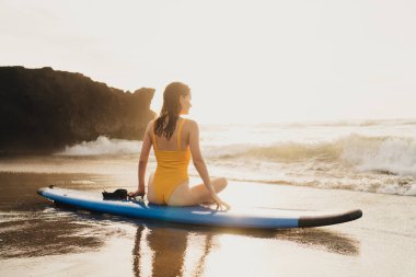 Yazın kumlu sahilden gelen fırtınalı okyanusa bakarken mayo giymiş, sörf tahtasında dinlenen tanınmamış bir kadın sörfçünün arka görüntüsü.