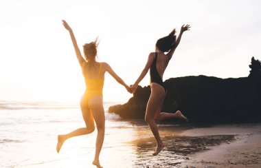 Anonim kız arkadaşların el ele tutuşup kumlu sahilde okyanus ve uçurumun dalgalı sularına atladığı arka plan.