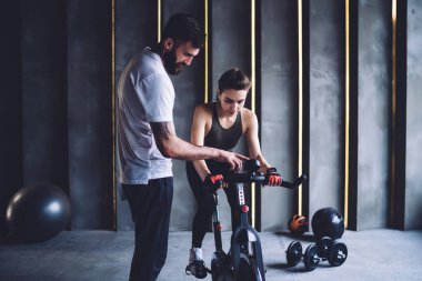 Yakışıklı erkek spor hocası bayan müşterisine zayıflamak için egzersiz bisikletini nasıl kullanacağını gösteriyor. Birbirlerine aşık çiftler spor salonunda sağlıklı yaşam tarzlarını sürdürmekten memnunlar.