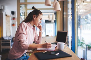 Düşünceli uzaktan kumandalı kadın çalışanın gündelik giysiler içinde masaya oturmuş açık not defteri ve dizüstü bilgisayarla notlar alırken görüntüsü.