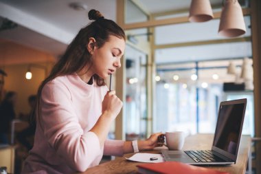 Yeni bir proje üzerinde çalışan genç kız öğrencinin yan görünüşü. Laptopuyla masada oturması ve kafede bir fincan sıcak içecek içmesi.